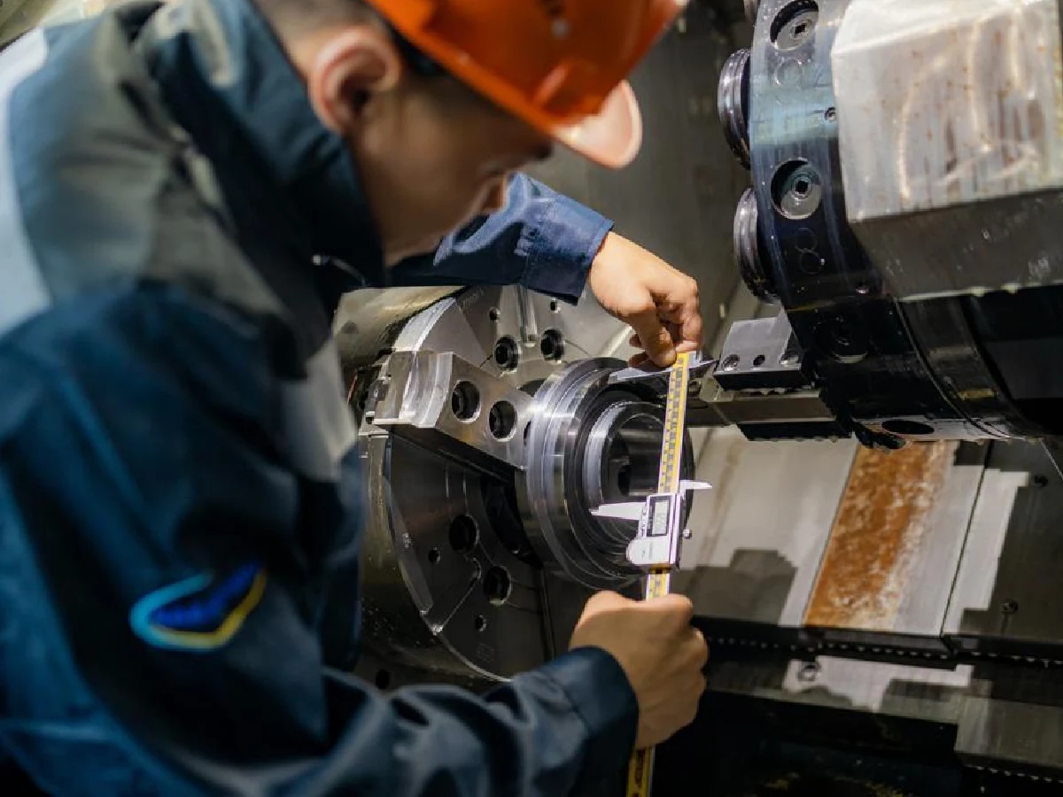 cnc-turning-operator-caliper-on-site-dimension-check - likundacnc.com 车间操作工佩戴安全帽,用数显卡尺现场测量数控车床加工金属工件外径尺寸